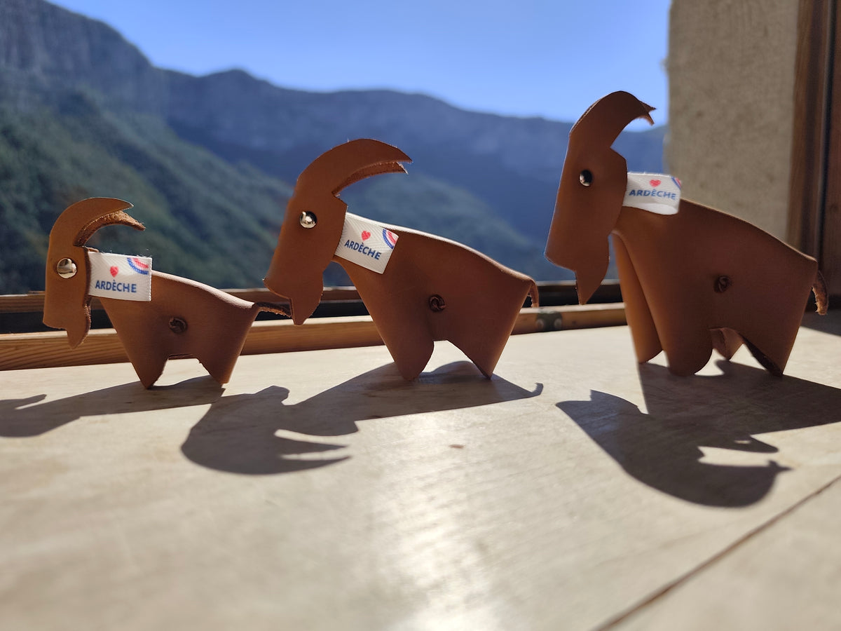 Petite, moyenne et grande chèvres en cuir camel avec étiquettes Ardèche, devant un paysage de montagne