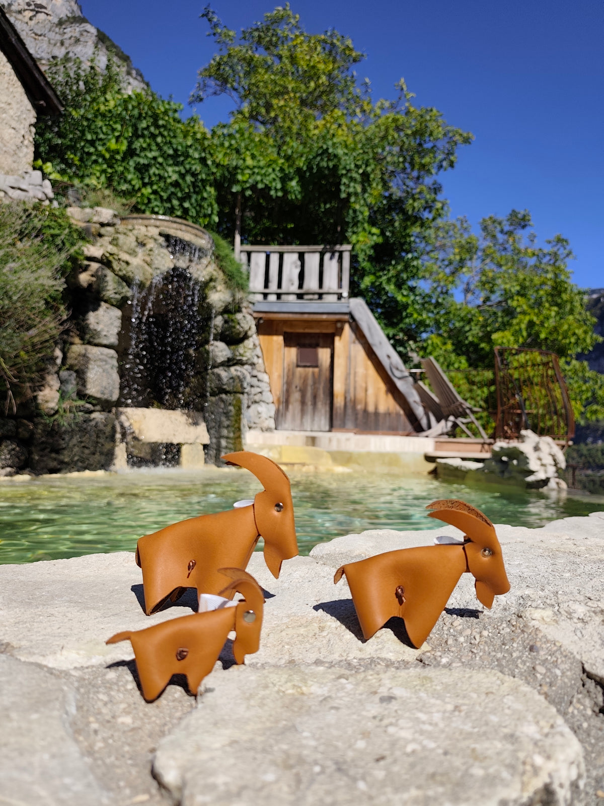 Trois chèvres décoratives en cuir camel de tailles différentes, sur un muret devant une fontaine