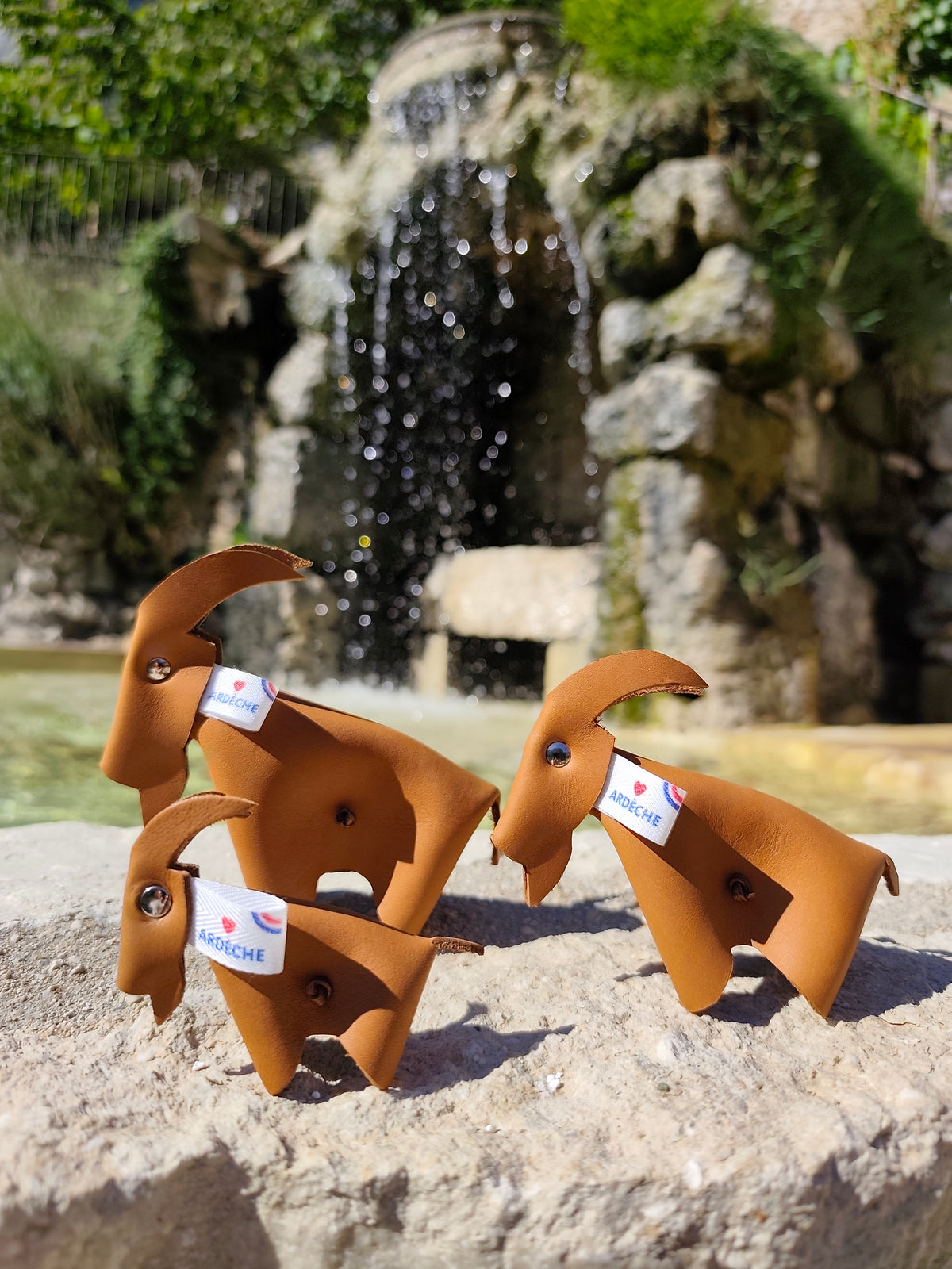 Trio de chèvres décoratives en cuir camel avec étiquettes Ardèche, devant une fontaine